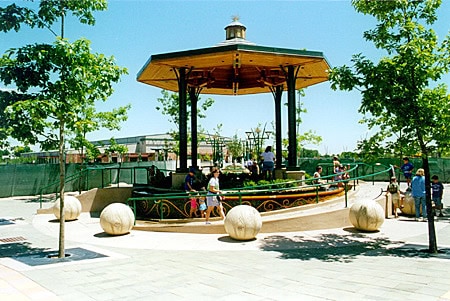 Octagonal wooden bandstand (gazebo) shelters people seated and standing; located on a sunlit urban plaza with young trees, four large spherical stone bollards, low decorative railing and buildings behind.