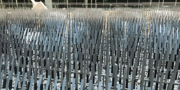 Rows of slender vertical metal slats rise in a dense, repeating grid, creating a rhythmic pattern above a bed of gray river stones inside a bright, glass-walled atrium.