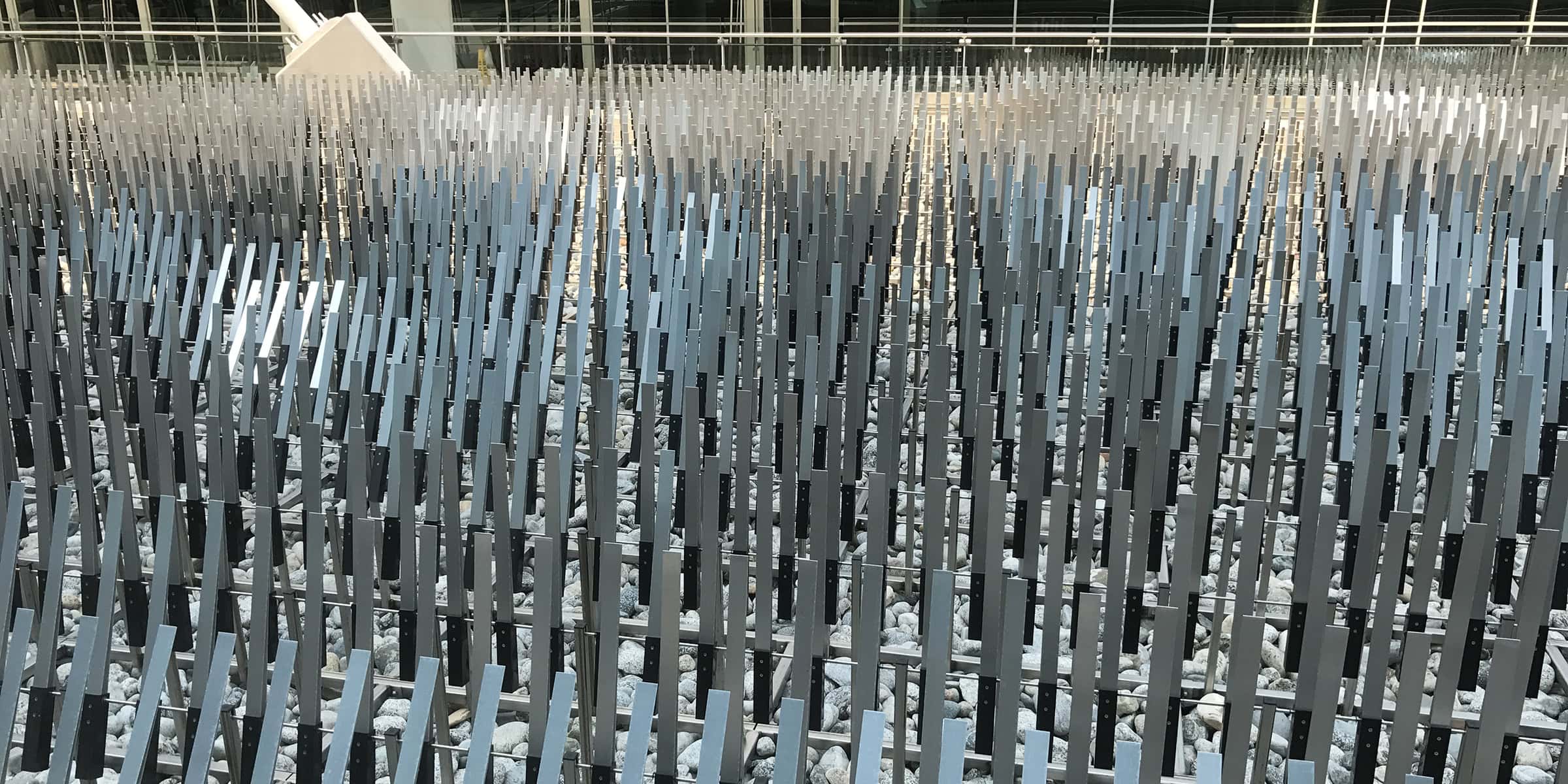 Rows of slender vertical metal slats rise in a dense, repeating grid, creating a rhythmic pattern above a bed of gray river stones inside a bright, glass-walled atrium.