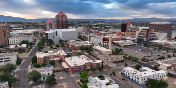 Downtown buildings cluster beneath darkening clouds, filling city blocks with mid-rise offices, parking lots, tree-lined streets, and distant mountains on the horizon.