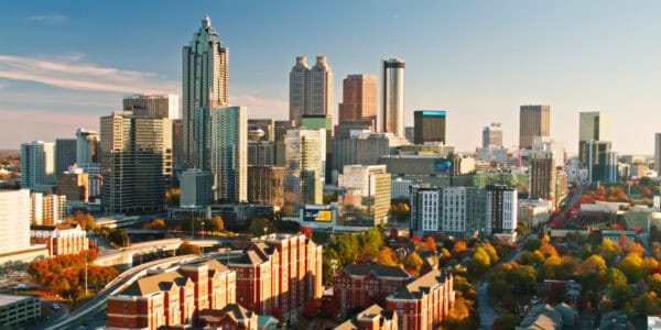 A cluster of glass-and-steel skyscrapers rises and reflects late sunlight over downtown, framed by highways and red-brick residential buildings in foreground and colorful autumn trees throughout.