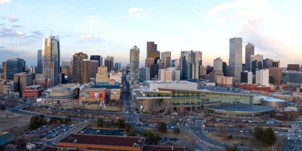 Glass-and-concrete skyscrapers tower over a low-slung convention center while cars travel along intersecting roads and parking lots in the foreground, under a pale evening sky.