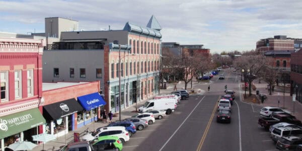 Three-story brick storefronts line a downtown street, hosting pedestrians and parked cars while vehicles drive past under bare trees and a cloudy sky.

Text visible: "HAPPY LUCKY'S TEAHOUSE"; "Curiosities"; "STOP"