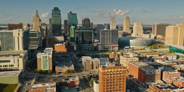 A cluster of downtown high-rise buildings rises above intersecting streets and low brick blocks, sunlight casting long shadows; a circular arena sits to the right in an aerial mid‑day view.

Visible text: "3"; "PRESIDENT"; "TWO LIGHT"; "EMERY BIRD THAYER CO. PRIVATE WAREHOUSE"; "T-Mobile Center"