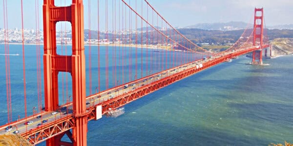 Golden Gate Bridge stretches across the bay, supported by tall orange-red towers and suspension cables, carrying vehicles along its roadway between forested headlands and a distant city.
