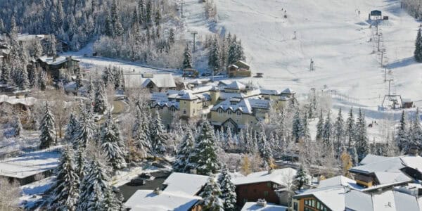 Clustered chalet buildings sit blanketed in fresh snow; a chairlift ascends powdery slopes; snow-dusted evergreens, a winding road and parked cars surround the mountain ski village.