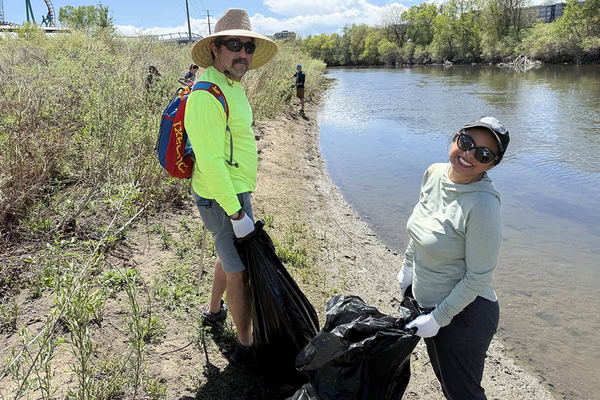 Two volunteers hold black trash bags, collecting litter along a sunny riverbank with low shrubs and calm water; one wears a wide hat and neon shirt, the other smiles toward the camera.

Text visible on red backpack (yellow letters): "maxi" (partly obscured).