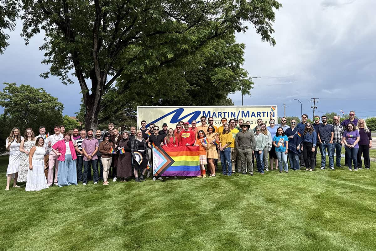 A diverse group poses holding a Progress Pride flag and rainbow flags in front of a "MARTIN / MARTIN" sign on grassy lawn under a large tree and cloudy sky.