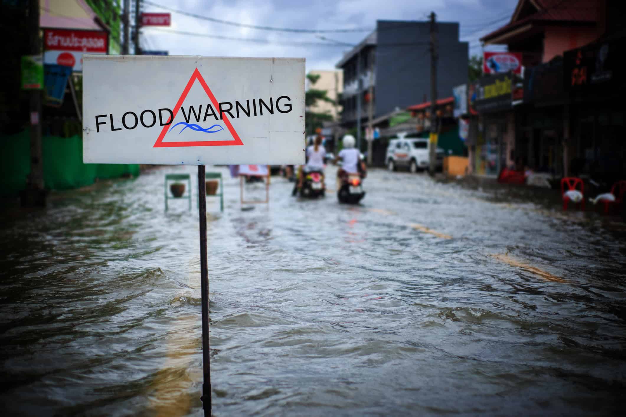 Road sign warns of flooding; it stands on a pole in rippling, knee-to-waist-deep water while motorcyclists travel down a waterlogged urban street lined with shops. 
Text: "FLOOD WARNING"