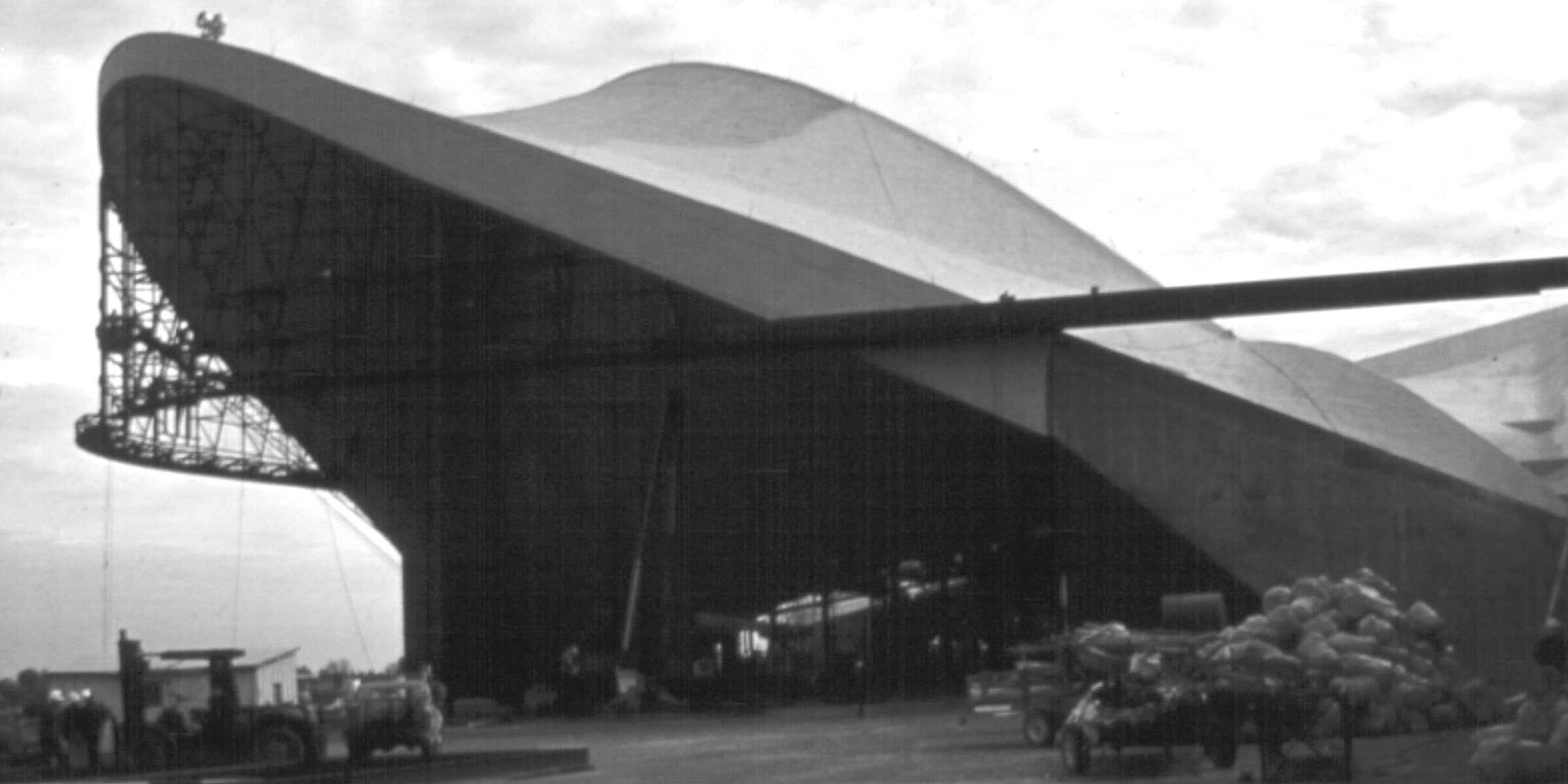 Massive curved hangar opens, revealing internal scaffolding; vehicles and piled cargo sit on a tarmac in front under a cloudy sky.