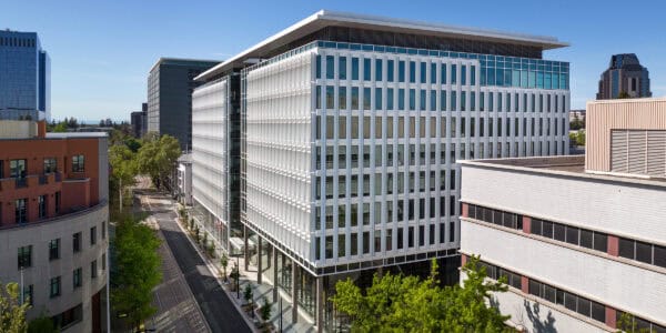 A white-glass office building occupies a city block corner, rising above a tree-lined street with tram tracks, flanked by neighboring mid-rise buildings under a clear blue sky.