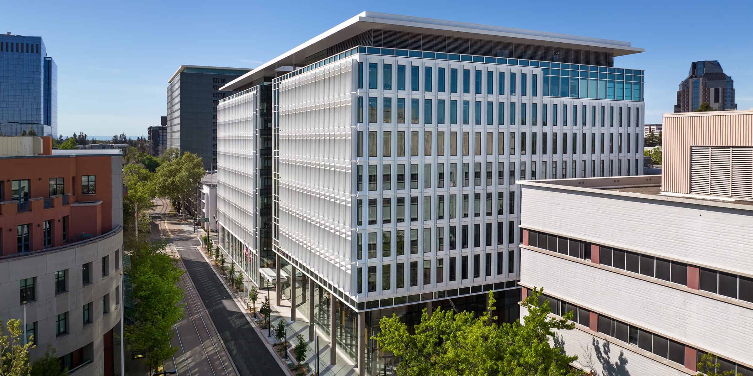 A white-glass office building occupies a city block corner, rising above a tree-lined street with tram tracks, flanked by neighboring mid-rise buildings under a clear blue sky.