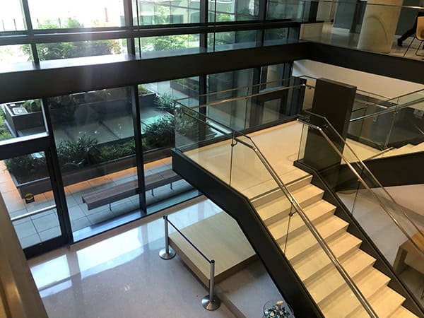 A glass-and-metal staircase ascends from a polished lobby floor to an upper mezzanine, flanked by glass railings, large floor-to-ceiling windows, planter beds and a wooden bench outside the atrium.