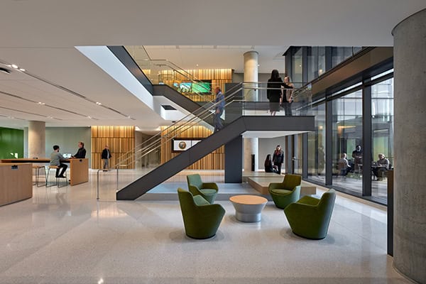Open central staircase: people ascend and descend between floors in a modern glass-walled atrium with polished terrazzo floors, green lounge chairs clustered around low tables, reception desks and wood accents.