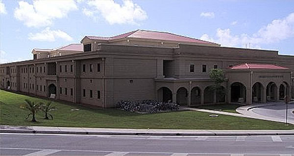 Large two-story beige institutional building with red roofs sits on a grassy lawn; arched entrances and a bicycle rack near the main entrance face a curved driveway and blue sky.
