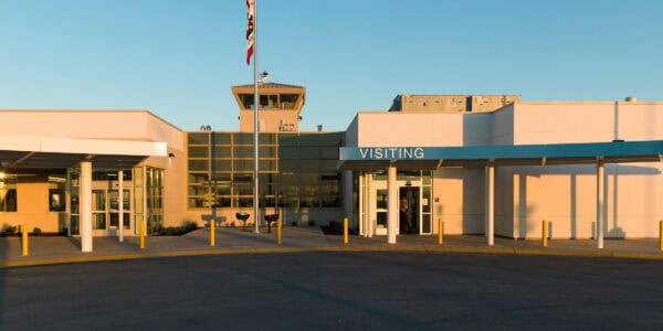 Entrance canopy labeled "VISITING" shelters a person entering through glass doors, framed by a flagpole and control tower above, set in a low modern facility with an empty drop-off circle at sunset.