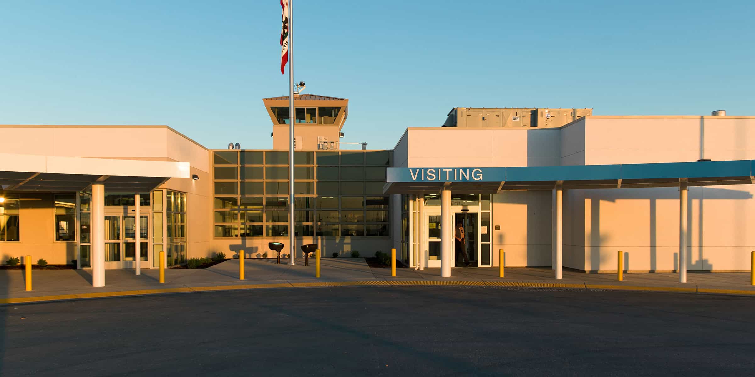 Entrance canopy labeled "VISITING" shelters a person entering through glass doors, framed by a flagpole and control tower above, set in a low modern facility with an empty drop-off circle at sunset.