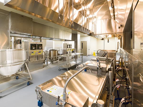 Stainless-steel commercial kitchen equipment lined in a production line, connected and ready for use, occupying a bright, sterile industrial kitchen with large ventilation hoods, sinks, mixers and wheeled kettles.