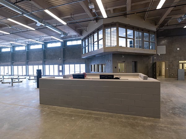 Concrete-block reception desk with monitors and a seated worker; set inside a spacious industrial hall with concrete floors, high ceiling, large windows and an elevated glass observation booth.