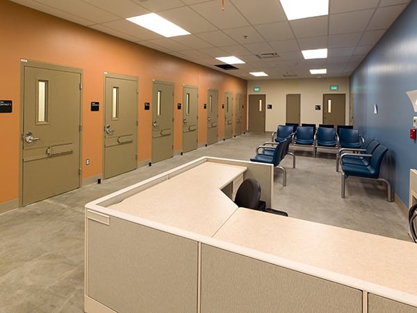 Reception desk sits empty, facing a row of closed beige doors; blue chairs line the opposite wall in a hallway with orange-blue walls and concrete floor. "EXIT" signs above doors.