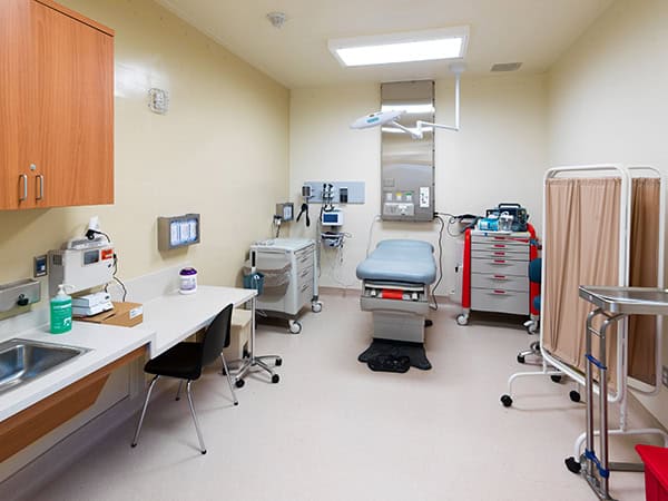 Exam table — centered, prepared for patient — clinical exam room with overhead light, monitors and vital‑sign equipment, supply cabinets, sink, rolling carts, privacy curtain and stainless instrument tray.