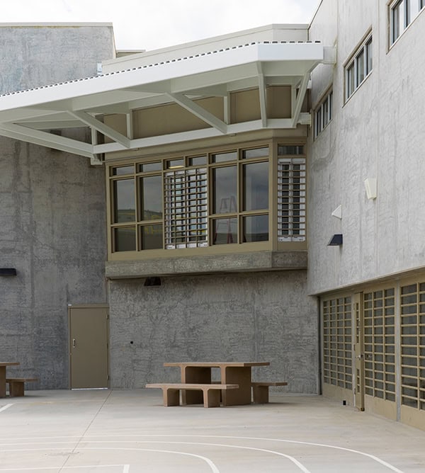 Concrete picnic table sits centered on a paved courtyard, beneath a white steel canopy and adjacent to tall gray concrete walls with multi-pane windows and a closed metal door.