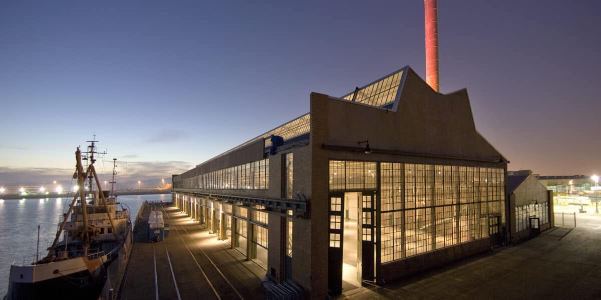 Industrial waterfront warehouse glows with interior lights as a docked cargo ship sits alongside, while a tall red-lit smokestack rises above against a twilight harbor with distant lights.