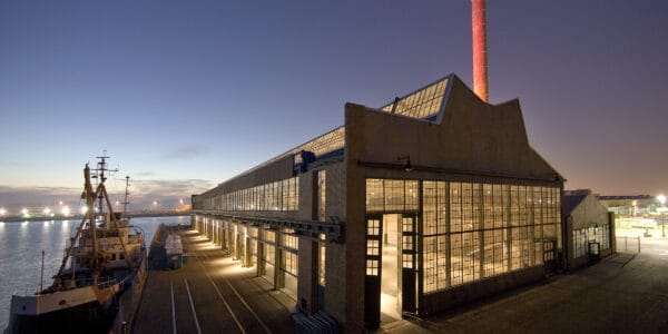 Industrial waterfront warehouse glows with interior lights as a docked cargo ship sits alongside, while a tall red-lit smokestack rises above against a twilight harbor with distant lights.