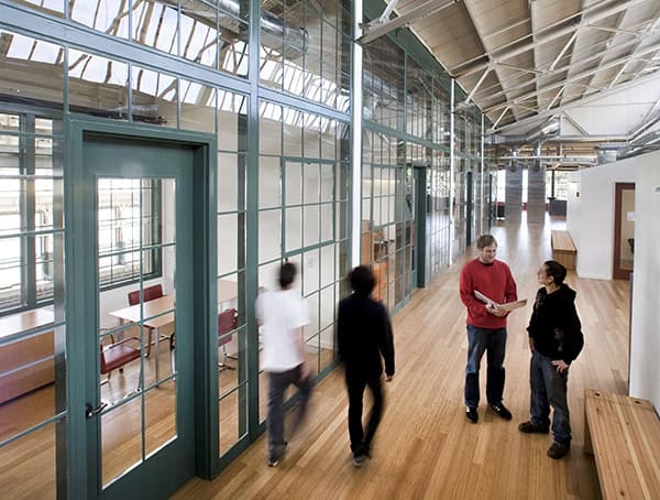 Glass-walled office corridor frames two people talking while two others walk past, inside a high-ceiling, industrial-style workspace with wooden floors, benches, and glass-partitioned meeting rooms.