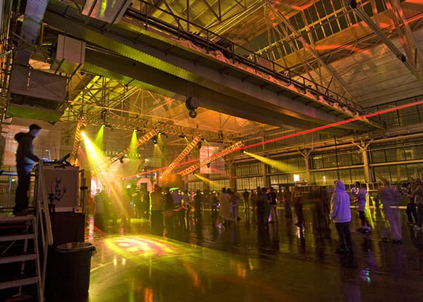 Large crowd gathers, milling and dancing under colorful stage lights and laser beams, inside a spacious industrial warehouse with exposed steel trusses, high ceiling, and a glossy concrete floor.