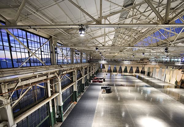 Large industrial hangar sits mostly empty, lit by overhead lights; a few seating clusters rest on polished floor beneath exposed steel trusses and tall multi-pane windows showing dusk sky.
