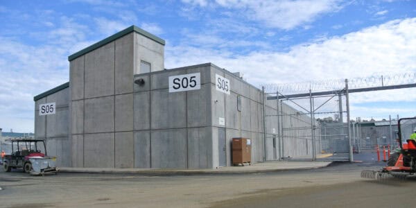 Concrete security building labeled "S05" sits inside a fenced compound topped with razor wire; a utility vehicle and a tractor work on the paved yard under a blue, partly cloudy sky.

Text transcribed: "S05" (appears on three panels of the building)