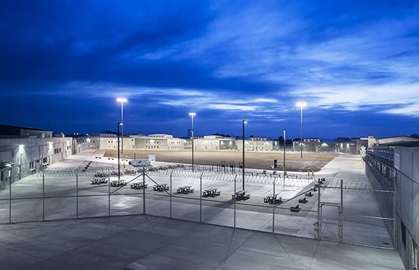 Prison yard sits empty, brightly lit by tall floodlights; picnic tables arranged inside chain-link and barbed-wire fencing, surrounded by low beige cellblocks beneath a deep blue evening sky.