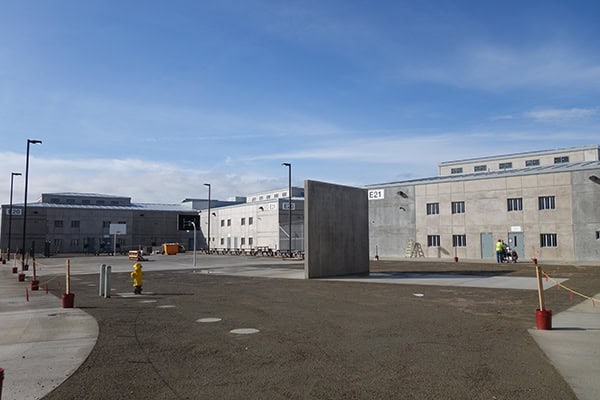 Concrete freestanding slab labeled "E21" stands in an expansive asphalt courtyard; surrounding low concrete buildings marked "E20", lampposts, a yellow fire hydrant and two workers under blue sky.

Text transcribed: "E21", "E20"