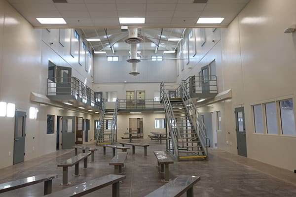 Metal dining benches and twin metal staircases dominate a spacious, high-ceilinged common room. Benches sit in rows; staircases ascend to a second-level walkway flanked by small windows and beige walls.