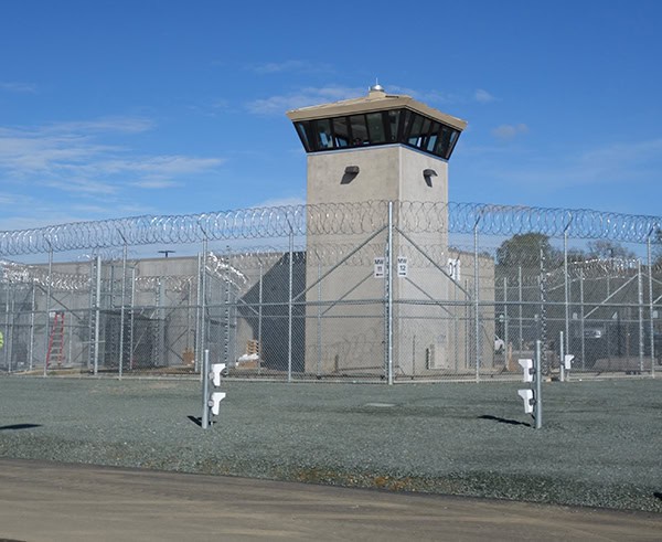 Watchtower monitors a secured prison yard, standing within barbed-wire-topped chain-link fences; gravel foreground and clear blue sky frame the perimeter.