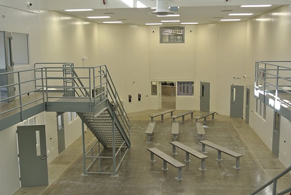 A concrete institutional dayroom with rows of bolted metal benches; a central steel staircase and mezzanine; closed gray cell doors and small windows line high‑ceilinged, fluorescent‑lit walls.