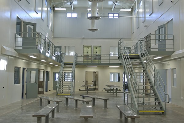 A two-tier cellblock with metal benches sits empty; twin staircases ascend to grated second-level walkways surrounded by small doors and windows in a high-ceilinged institutional room.