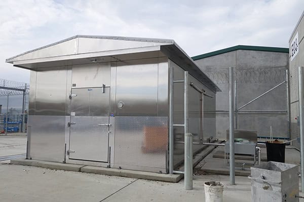 Stainless-steel utility shed with double doors and diamond-plate lower panels sits on a concrete pad, flanked by chain-link fencing topped with barbed wire, amid concrete industrial buildings under an overcast sky.