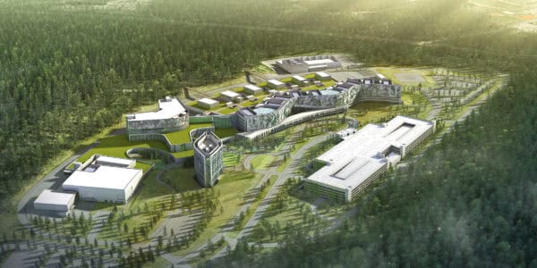 A sprawling modern campus complex stretches across a grassy clearing, its sinuous multi-wing buildings with green roofs connected by roads and walkways, surrounded by dense pine forest and parking structures.