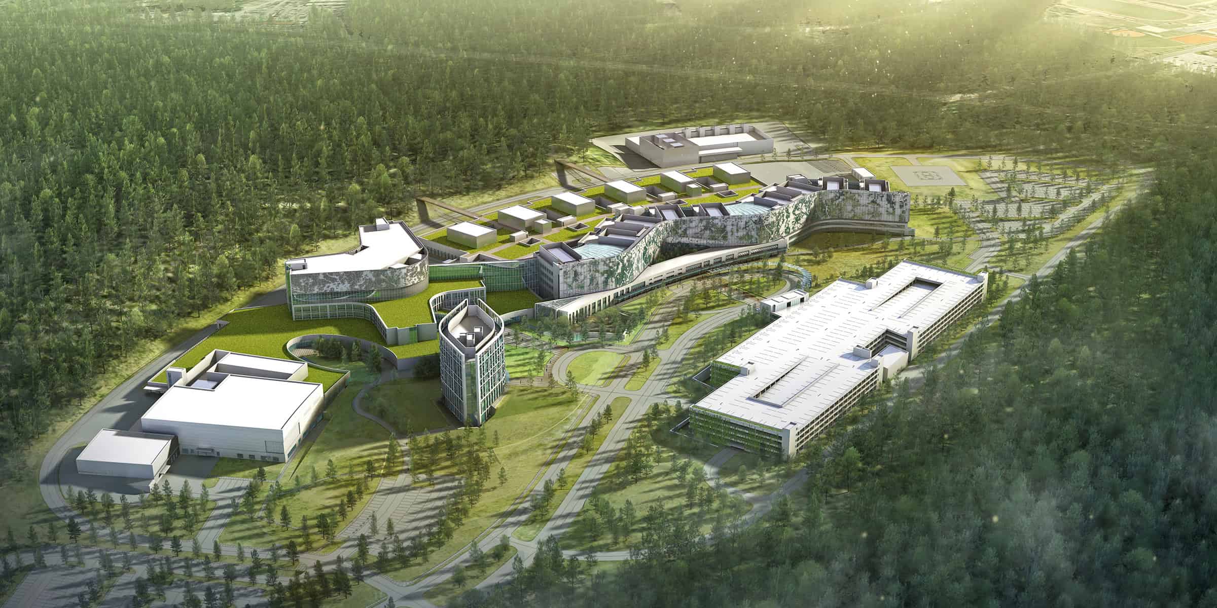 A sprawling modern campus complex stretches across a grassy clearing, its sinuous multi-wing buildings with green roofs connected by roads and walkways, surrounded by dense pine forest and parking structures.
