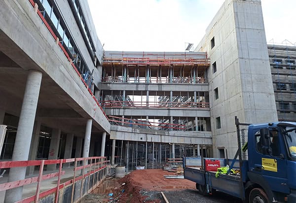 Multi-story concrete building under construction, with scaffolding and temporary supports spanning open floors; a flatbed truck and construction materials sit on muddy ground beside red "HILTI" toolbox, overcast sky above.