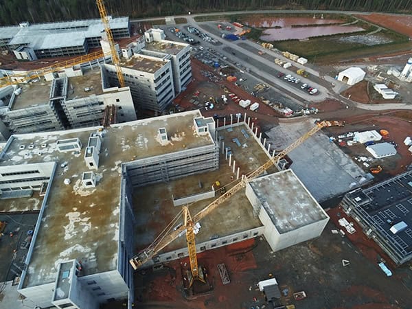 Large concrete building complex under construction with two yellow tower cranes lifting materials over an inner courtyard; muddy roads, scattered equipment and parked vehicles surround the site near a forest.