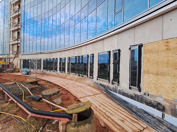 Curved glass building facade with narrow vertical windows is under construction; lower panels are being installed. Temporary wooden walkway, exposed red soil, cables, open manholes and scaffolding surround the site.