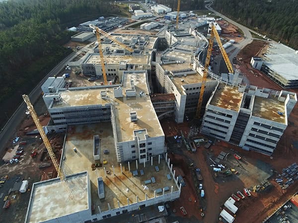 Concrete multi-wing building complex under construction, with several tower cranes, exposed floors, scaffolding, and construction vehicles on a cleared red-earth site surrounded by forest and access roads.