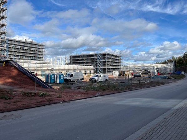 Scaffolded multi-story buildings rise behind parked vans and blue portable toilets on a muddy construction lot beside a paved road, scattered equipment and materials under a partly cloudy sky.