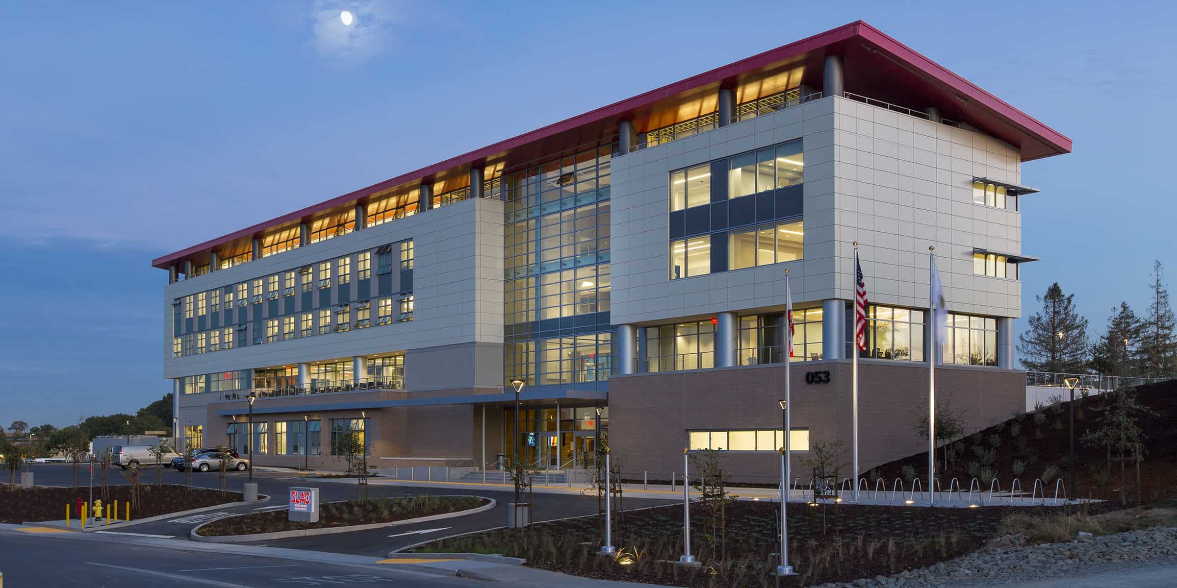 Modern multi-story office building glows with interior lights, facing a curved driveway and flagpoles at dusk; landscaped beds and parked cars surround the glass-and-panel façade. Text visible: "053", "SLAC".