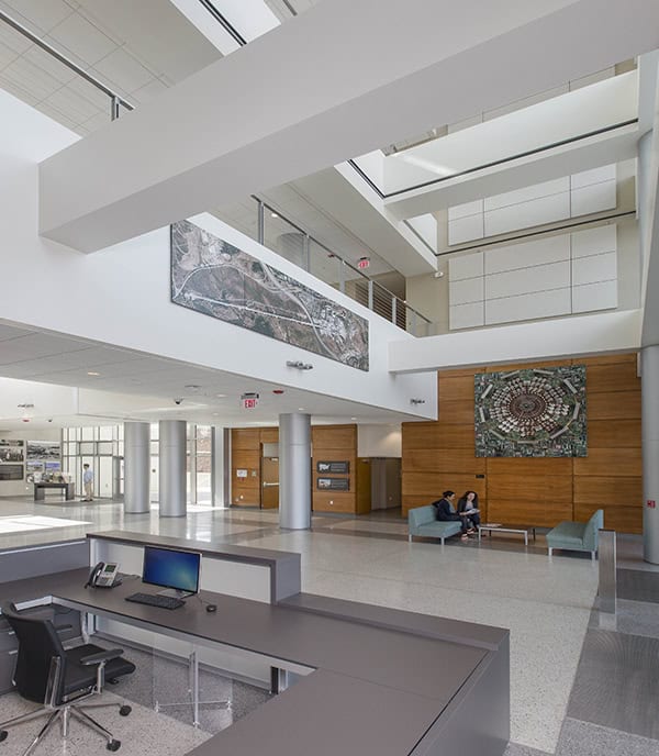Reception desk anchors the foreground, empty with a computer; two people converse on sofas by a wood-paneled wall; expansive two-story atrium lit by skylights with large wall murals. Visible text: "EXIT".