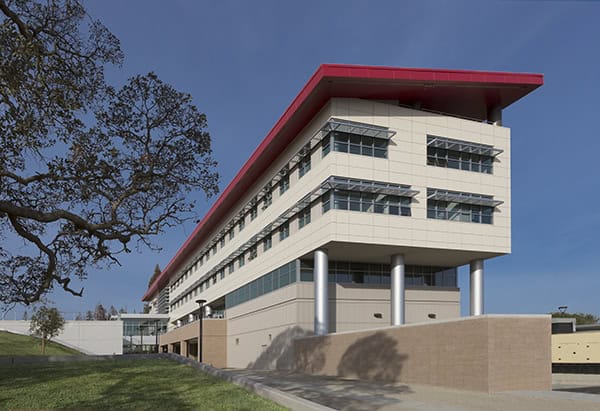 Contemporary multistory building with red overhanging roof, projecting balconies, and cylindrical columns supporting an upper corner; sits beside a grassy slope and large tree under a clear blue sky.
