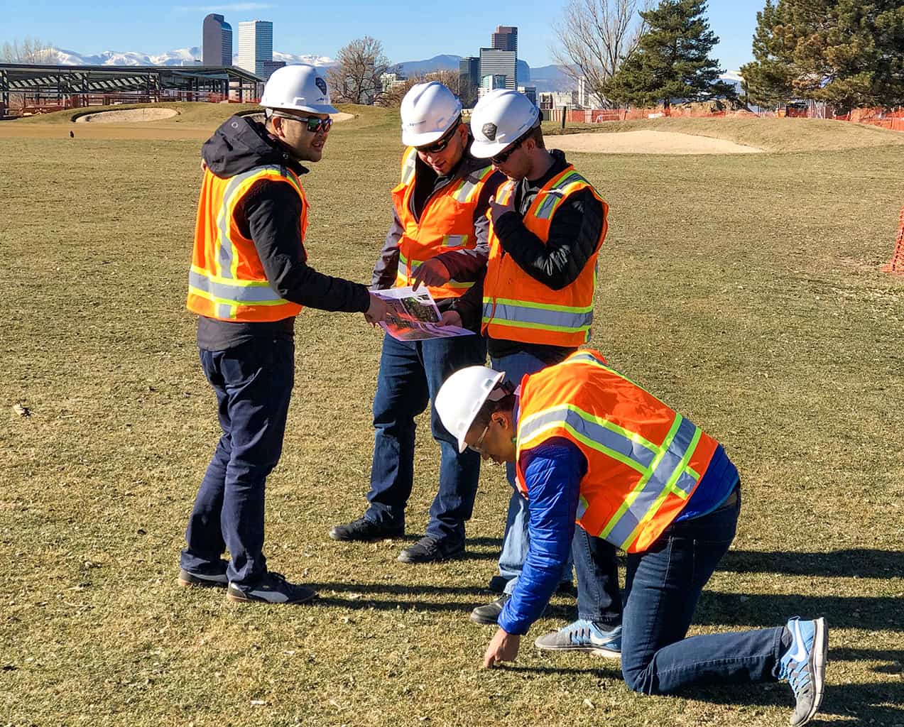 Four workers in white hard hats and orange vests study plans; one kneels and points at the ground on a grassy field with a city skyline and snow-capped mountains beyond.